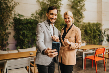Fototapeta premium Two smiling professionals take a selfie at an outdoor cafe. They share a moment of partnership and casual business style.