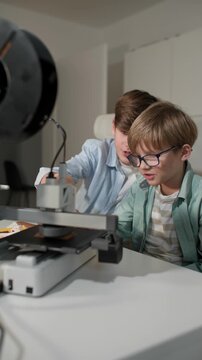 At a bright table, two schoolchildren watch a 3D printer work and control it