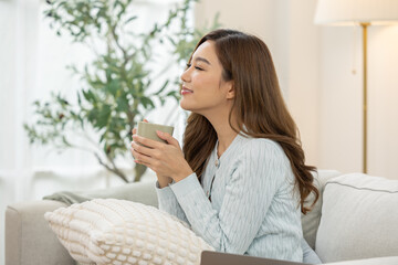 Happy relaxed young Asian woman holding hot coffee cup smelling aroma sitting on comfortable sofa...