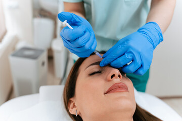 Fototapeta premium Practitioner in blue gloves performing a cosmetic botulinum toxin injection on a smiling woman's forehead during a professional aesthetic treatment in a medical spa setting.