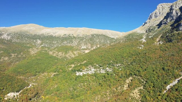 Aerial view of Papingo village nestled among lush green mountains under a clear blue sky in Greece. Picturesque traditional stone houses surrounded by dense forest and dramatic rocky peaks.