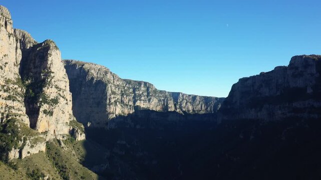 Dramatic limestone cliffs of Vikos Gorge rise steeply under a clear blue sky, casting deep shadows across the valley in northern Greece.