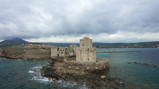 Aerial view of the historic Methoni Castle on the rocky coastline with turquoise sea and dramatic cloudy sky in Greece. The ancient fortress stands prominently by the water, surrounded by natural