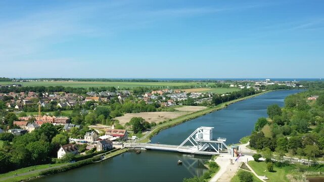 Scenic aerial view of Pegasus Bridge spanning the Caen Canal, surrounded by lush greenery and the town of Benouville, Normandy, France, under a clear blue sky.