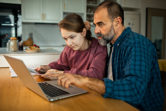 Father helping teenage daughter with homework on laptop at kitchen table