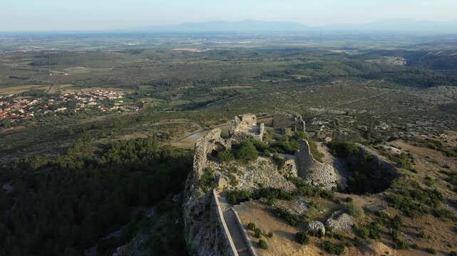 Drone shot of the ancient stone ruins of Chateau de Opoul Perillos surrounded by green hills and a distant village in southern France.