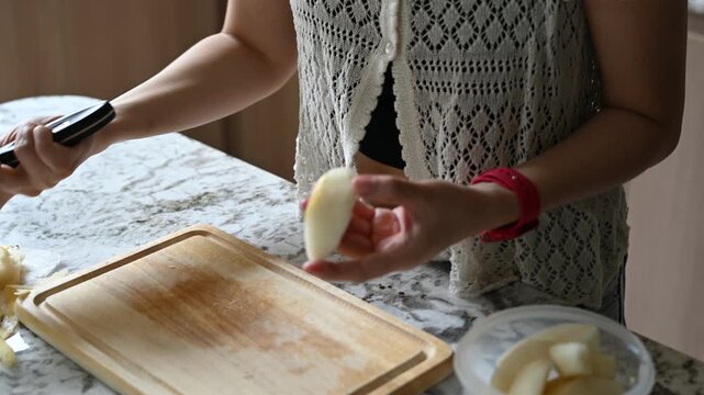 Asian woman quiet home lifestyle focused on relax and wellbeing. Healthy kitchen food preparation, peaceful moment peeling fresh pear for simple, homemade meal