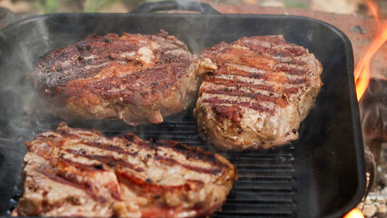 Three juicy steaks sizzling and smoking on a cast iron grill pan with an open fire