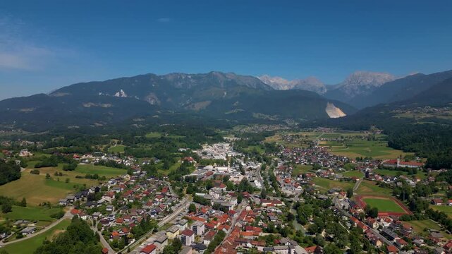 Wide aerial view of the historic town of Kamnik, Slovenia, featuring red-roofed houses, a central church, and a sports stadium, set against the towering peaks of the Kamnik-Savinja Alps in summer.