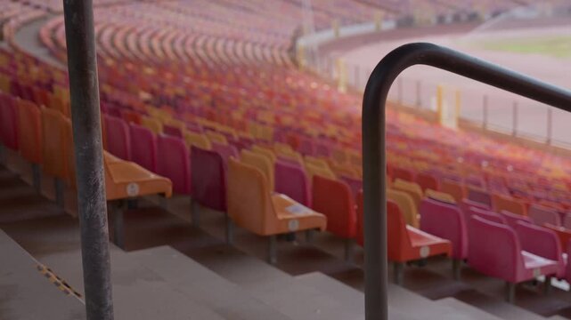 Empty colorful stadium seats with handrail, rows of orange and red plastic seats curve toward blurred playing field, concrete steps and metal railing in foreground, soft morning light casts muted