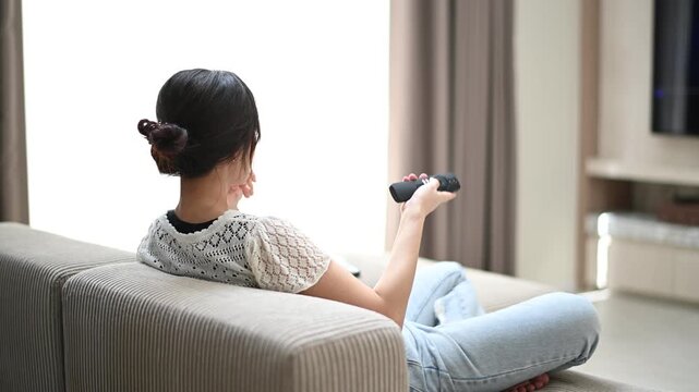 Calm asian woman promoting wellbeing with relaxing lifestyle at home. Enjoying quiet entertainment, watching television on sofa to relax in living room