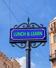 Fototapeta premium Lunch and learn symbol. Concept words Lunch and learn on beautiful big blue road sign. Beautiful blue sky and buildings background. Business, educational and lunch and learn concept. Copy space.