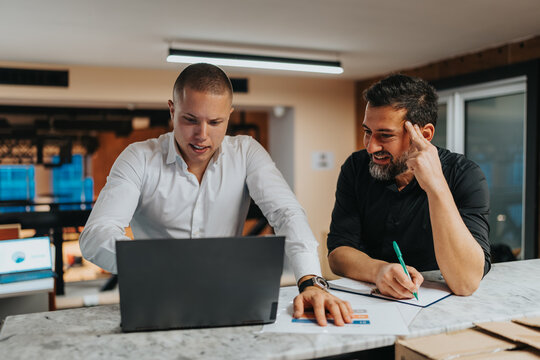 Two men collaborate at a modern office, one in white shirt explains something on a laptop. They review charts and notes together, showcasing teamwork and professional discussion.