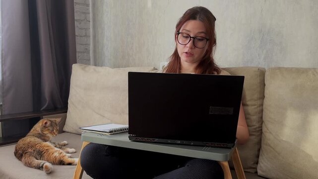 Young woman wearing glasses sitting on a sofa at home using a computer and talking animatedly during an online interview in a remote job recruitment setting