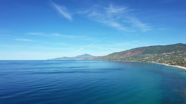 Scenic aerial view of the clear blue sea and rugged coastline of Palinuro, Italy, with mountains in the background under a bright sky.