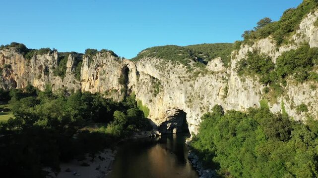 Scenic view of the Pont d Arc natural limestone arch spanning a river, surrounded by lush greenery and dramatic cliffs in Ardeche, France. Clear blue sky enhances the vibrant landscape.