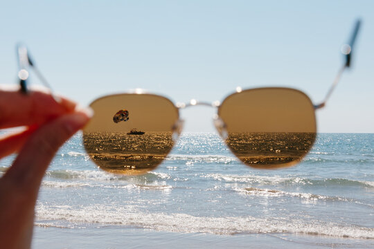 Hand holding sunglasses focusing on an ocean view, capturing a parasailer in the distance during a sunny day at the beach. Water sparkles under the clear blue sky. Summer travel creative concept.
