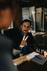 Fototapeta premium A man in a dark suit sits at a wooden table with a notebook and pen. He shows a peace sign with two fingers in a casual office setting.