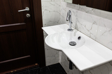 Fototapeta premium A modern bathroom sink with a sleek chrome faucet mounted on a white ceramic countertop. Set against marble-patterned tiles, next to a dark wooden door with a metallic handle.