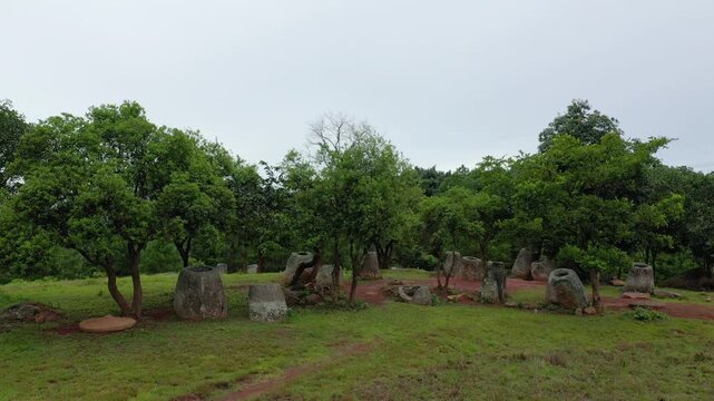 Large ancient stone jars scattered among green trees in the Plaine des Jarres, Laos, under an overcast sky. The mysterious landscape features archaeological relics in a rural setting.
