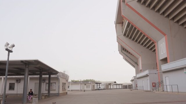 empty stadium exterior with lone person, security standing near canopy and ticket booths, wide concrete plaza bathed in soft dawn light, lamp posts casting long shadows, muted pastel sky, atmosphere