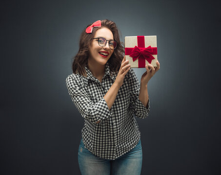 Surprised woman holding gift box - present with ribbon over dark gray - black background. retro style girl with a present at holiday Birthday.