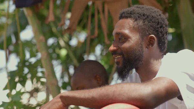 Black coach smiling with soccer ball in closeup profile under banana trees, expressive laughter and hands covering face gesture, candid mentor moment, sunlit beard and warm highlights, playful yet
