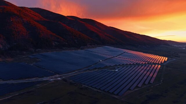 Large solar power plant under vibrant orange sunset sky. Wide aerial shot of photovoltaic station rows against dramatic red and orange sunset sky and mountain silhouette.