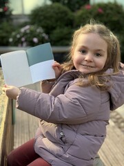 Smiling young girl holding an open blank greeting card outdoors. Child presenting empty paper card with copy space for text, message or announcement. Natural light, casual lifestyle concept.