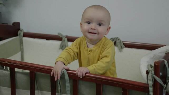 caucasian baby standing at crib smiling broadly, milestone achiever in yellow onesie showing joyful grin and playful gaze, bright nursery with potted plant and modern decor capturing