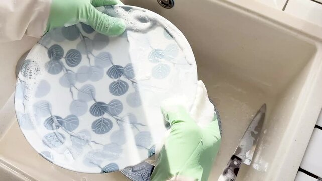 Man in green rubber gloves washes dishes in a kitchen sink using a sponge and liquid soap. Contemporary domestic scene and modern masculinity. Concept of sharing household chores. Day light, close-up