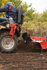 Obraz premium Man driving red walk-behind tractor, plow digging soil in garden, side view.