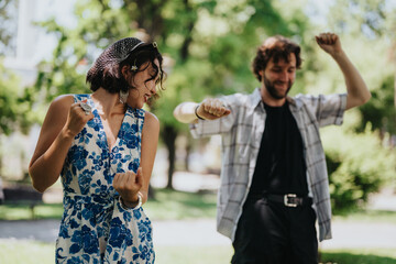 Fototapeta premium A woman in a blue floral dress dances with a man in a plaid shirt in a sunny park. They laugh and move together, enjoying a casual outdoor moment.