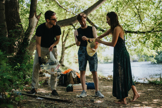 Three volunteers pick up trash along a shaded riverbank, wearing gloves and casual clothes. They work together outdoors to protect nature while bags of waste lie nearby.