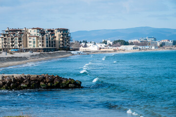 Obraz premium large, multi-story apartment building or hotel situated directly on the shoreline, with small waves breaking near a stone breakwater under a clear blue sky.