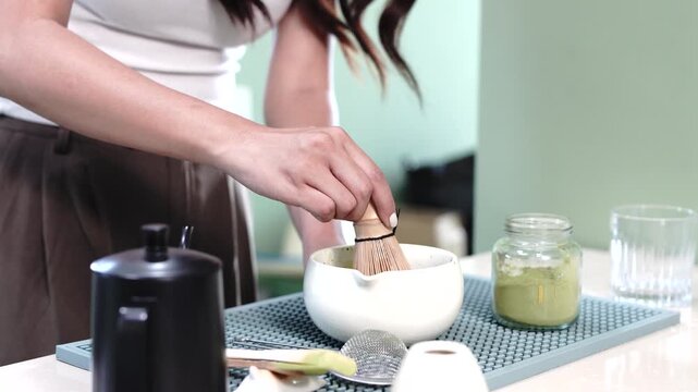 Asian woman prepares matcha tea by whisking green powder in a bowl with a bamboo whisk, showcasing the blending process in a bright kitchen setting