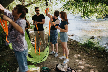 A group of friends stands by a river, preparing a bright orange strap and green fabric. They chat and set up equipment for a fun outdoor activity on a sunny day. © qunica.com
