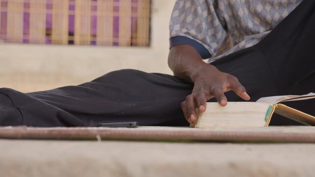 Black elder hands on book mat, weathered fingers turning pages beside phone, woven carpet texture, soft warm light, intimate domestic study, calm contemplative mood, focus on touch and tradition