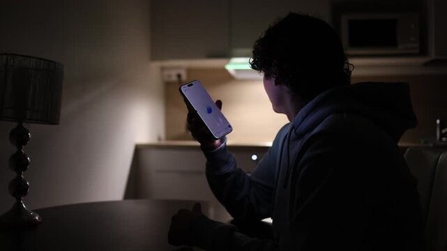 Young student at desk in dark apartment studying at night using smartphone communicating with chatgpt asking questions ai assistant helping with education and homework