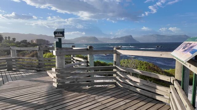 Simon's Town South Africa. 06.02.2026. Video. A wooden walkway with railings leads to a viewpoint offering expansive ocean vistas and hazy mountain range of Cape Peninsular under a dramatic sky