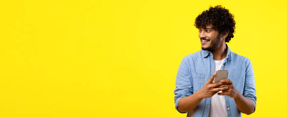 A young man stands and smiles while looking at his smartphone. He wears a casual blue shirt and has curly hair. The background is bright yellow, creating a vivid atmosphere. © Prostock-studio