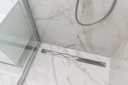 A modern shower area with marble-patterned tiles, linear stainless steel drain, glass door, and flexible chrome shower hose mounted on the wall.