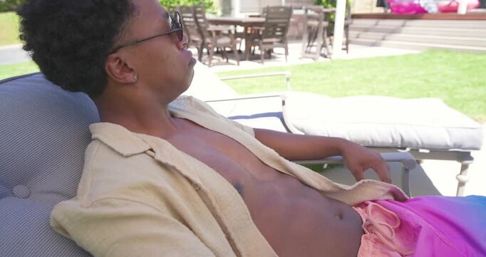 African man sipping stemmed glass, reclining on lounger in backyard, wearing shades, relaxing