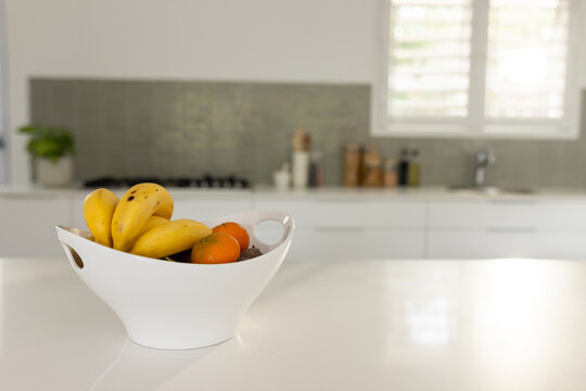 White ceramic fruit bowl is sitting on countertop near gas stovetop, holding bananas and oranges