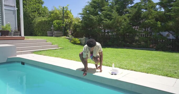 African American man in trunks walking poolside, pausing near hat and bottle, checking water temp