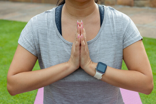 Kneeling female on pink yoga mat on grass, in gray top, pressing hands together, showing smartwatch