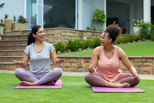 Diverse female friends sitting on purple yoga mats on backyard lawn, wearing leggings and chatting