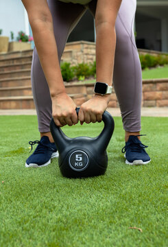 Woman in mid 30s holding 5 KG kettlebell, performing hip hinge on grassy lawn wearing smartwatch