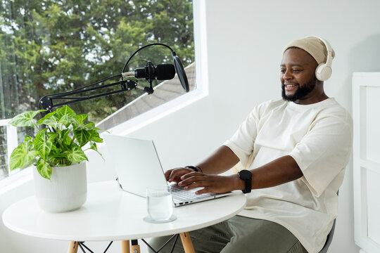 Adult African man typing on laptop in studio by window, beanie, headphones, microphone, copy space