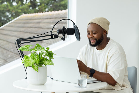 African man sitting at white table in bright studio, using laptop and microphone, wearing beanie
